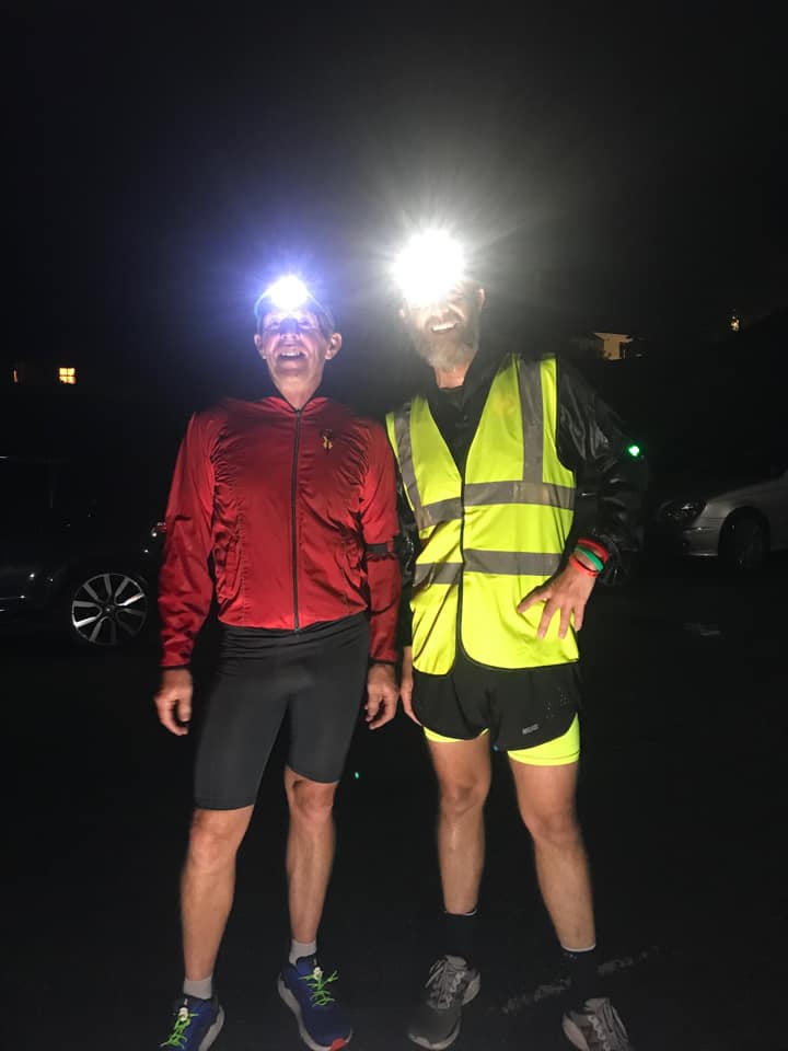 Ian dressed in a red running jacket and Chris in a high vis jacket pose for a photo smiling. They are both wearing head torches.