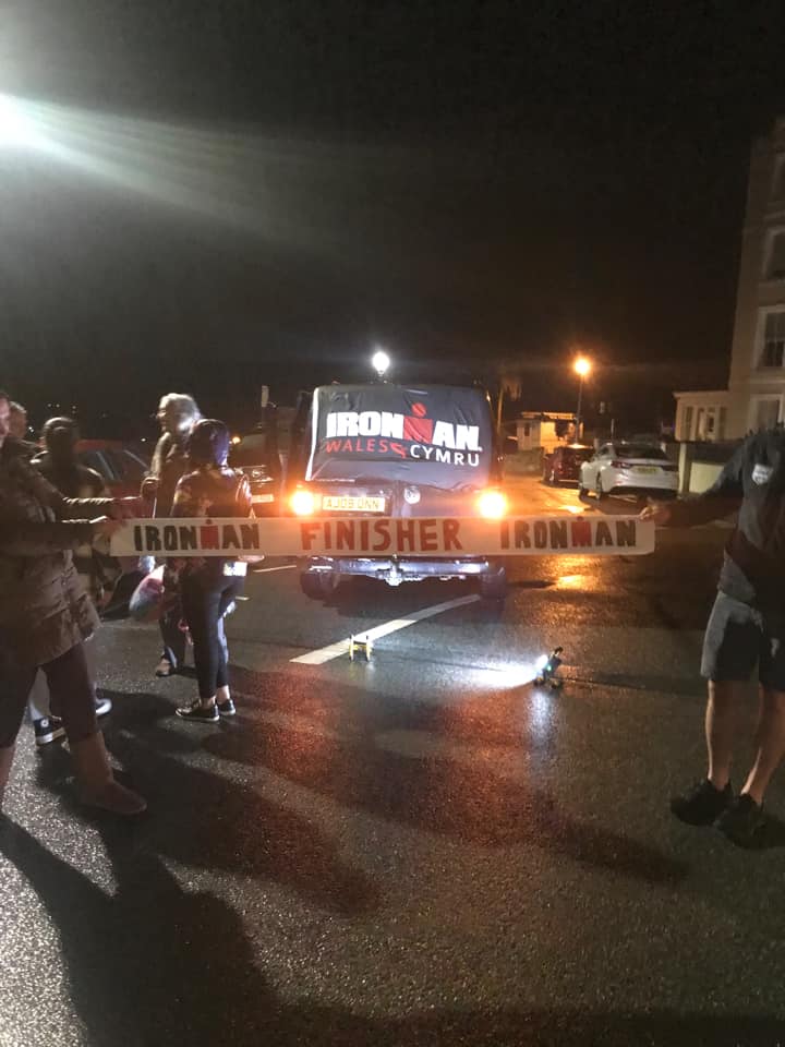 People holding up a homemade finish line banner which reads "Ironman finisher" there is an Ironman Wales flag just behind